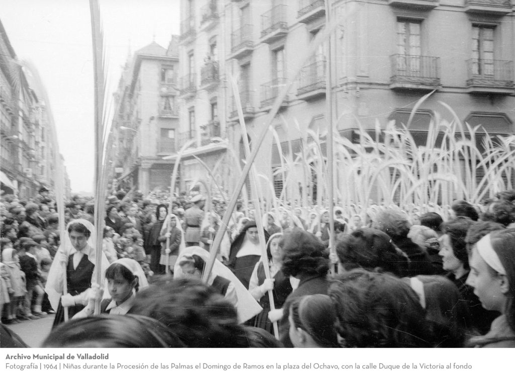 Fotografía. 1964. Niñas durante la Procesión de las Palmas el Domingo de Ramos en la plaza del Ochavo, con la calle Duque de la Victoria al fondo